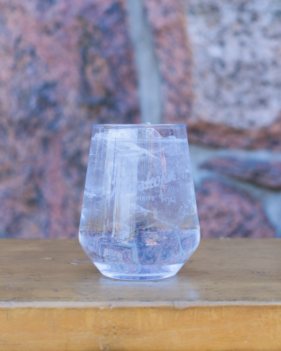 A clear stemless cocktail glass filled with ice, displayed on a wooden surface with a rock wall in the background.